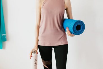 Faceless slim female athlete in sportswear standing with blue fitness mat and water bottle while preparing for indoors workout